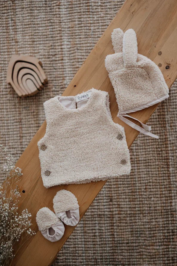 Driedelige teddyset voor baby's in beige, inclusief vest, muts en anit-slip slofjes, beschikbaar in maten 0-12 maanden en 12-24 maanden.