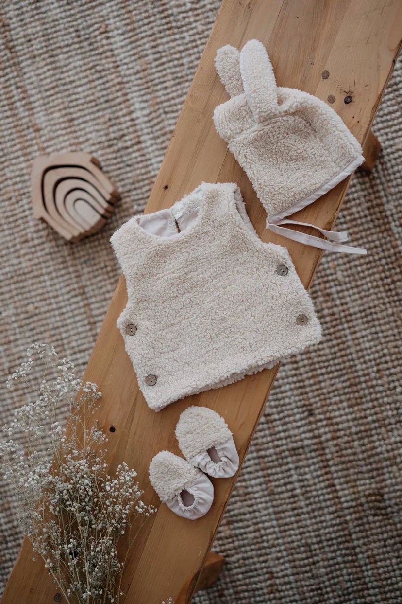 Driedelige teddyset voor baby's in ecru, inclusief vest, muts en anit-slip slofjes, beschikbaar in maten 0-12 maanden en 12-24 maanden.